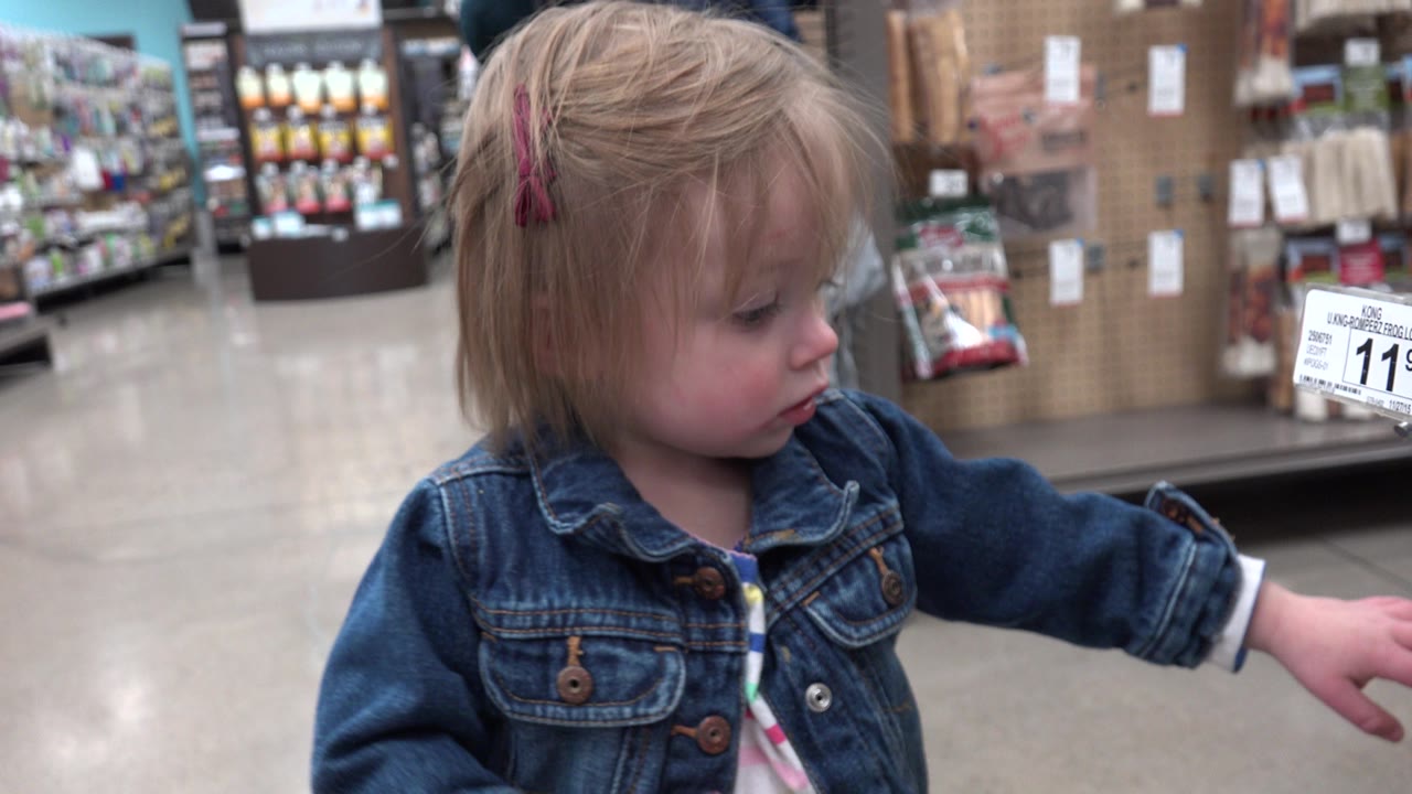 The young blonde girl remains in close foreground in the Denver supermarket, looking down while shelves, hanging products, and an 11.99 price sign fill the background.