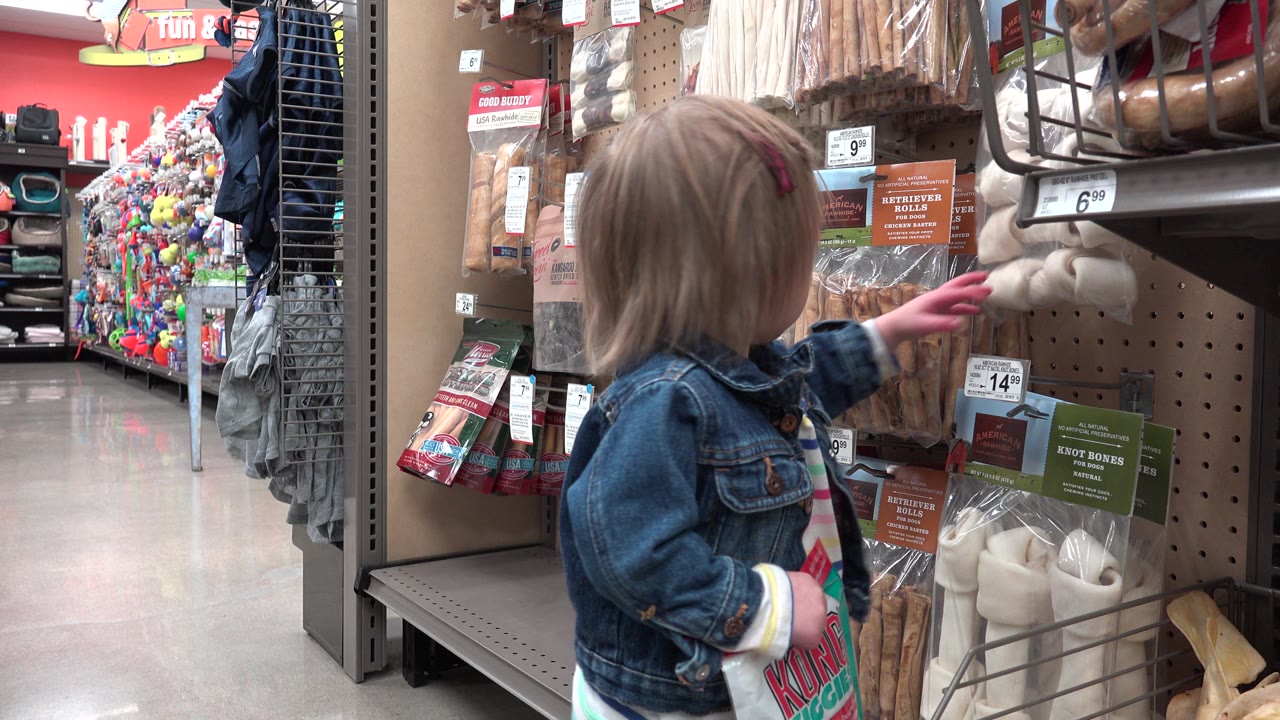 A young child touches a hanging package of pet chews while standing close to a pegboard display inside a Denver, Colorado store aisle.