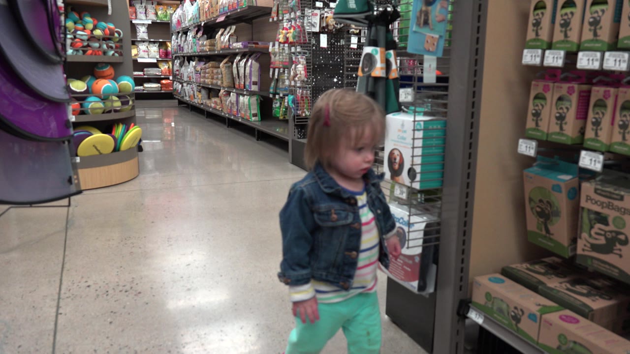 The toddler remains near the aisle end in Denver, facing shelves stocked with bright packaged items, balls, and display racks under indoor store lighting.