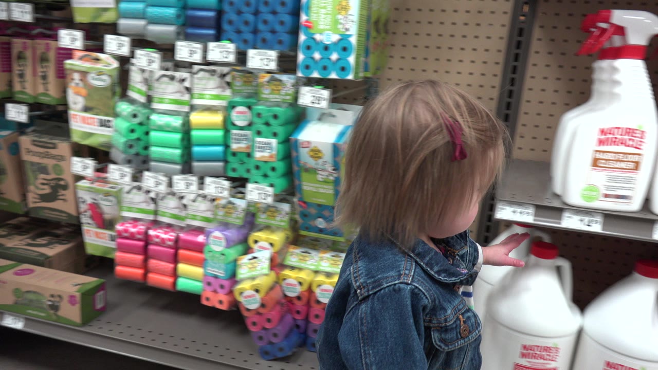 The child reaches one hand toward a large white jug on a lower shelf in the Denver retail aisle, with spray bottles and packaged products behind them.