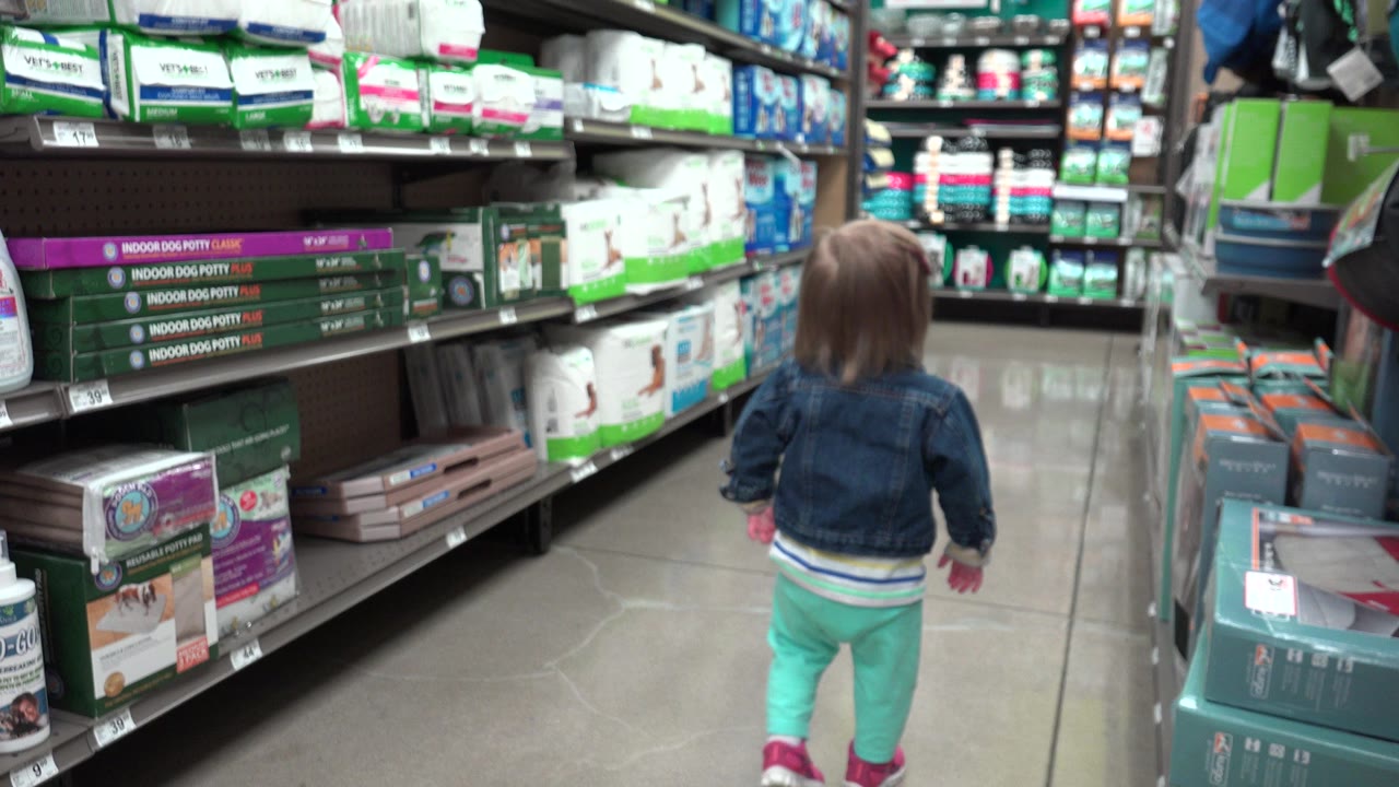 The child continues farther along the Denver retail aisle, seen from behind between shelves of boxed and packaged hygiene products.