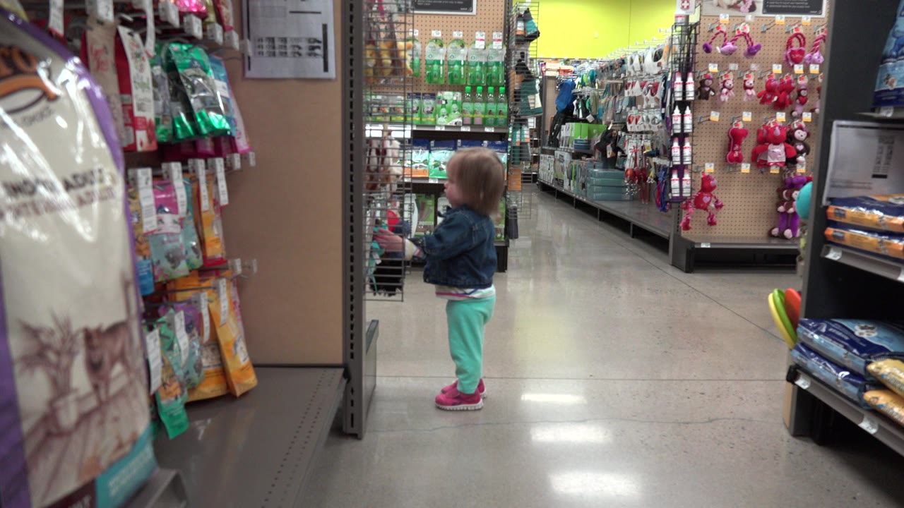 A toddler stands near an end cap display in a Denver, Colorado retail aisle, with hanging packages on the left and stocked shelves stretching into the background.