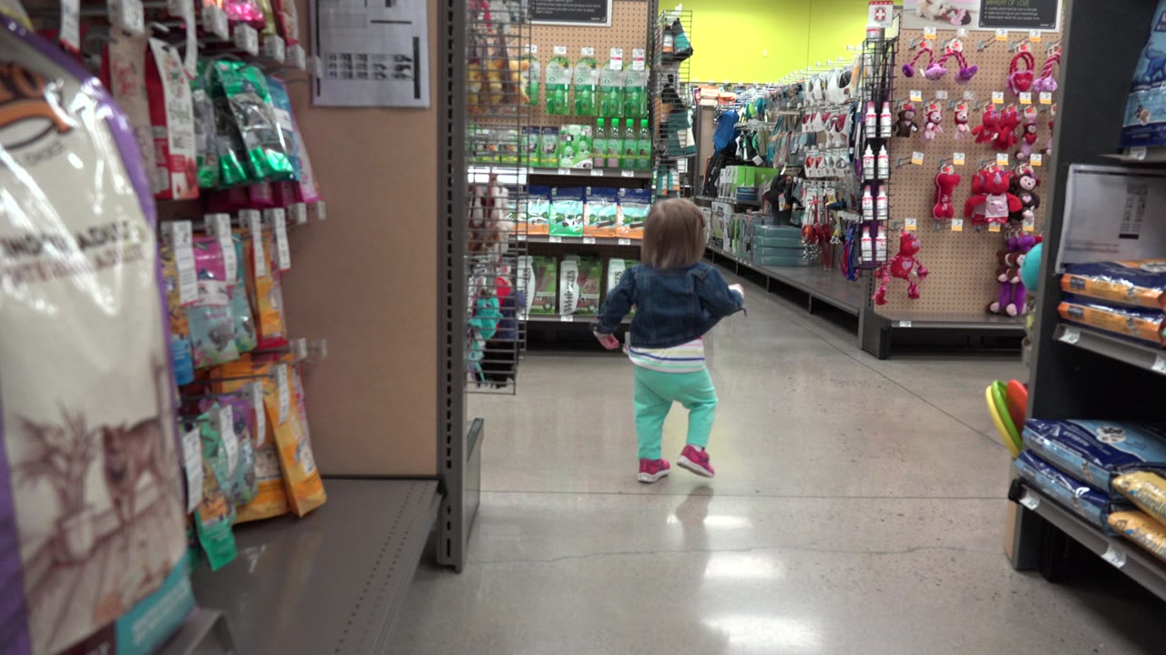A toddler girl in mint pants and a denim jacket walks away from the camera down a Denver retail store aisle lined with hanging products and shelves.