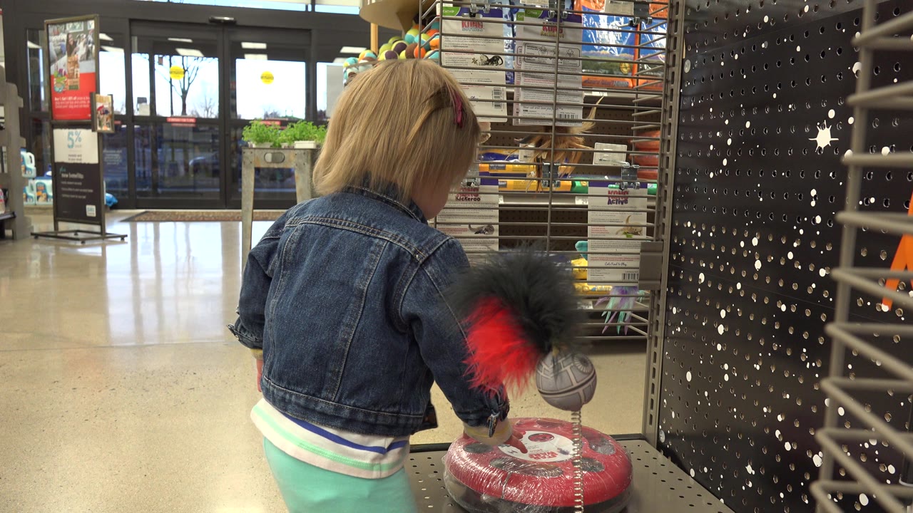 The child remains turned away from the camera in the Denver retail interior, with a red and black hanging toy and metal display hardware filling the right side.