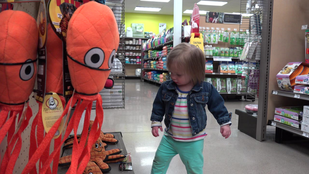 Young girl in Denver, Colorado stands near a tall orange plush toy display, with store shelves and fluorescent lighting behind her.