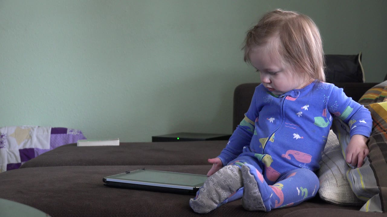 Young child sitting on a bed beside a tablet.