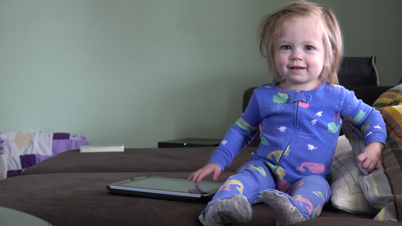 Toddler in blue pajamas sitting on a couch beside a tablet.