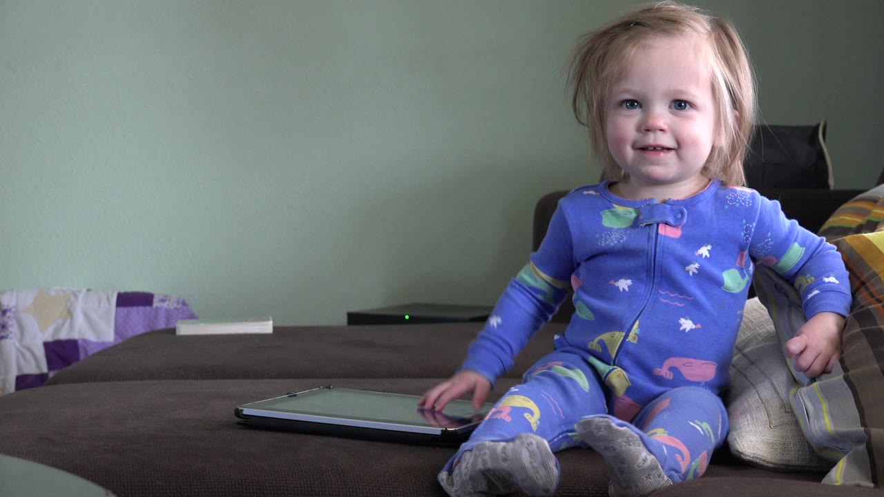 The child remains centered on the couch in Westminster, Colorado, with her hand closer to the tablet and pillows tucked beside her against the green wall.