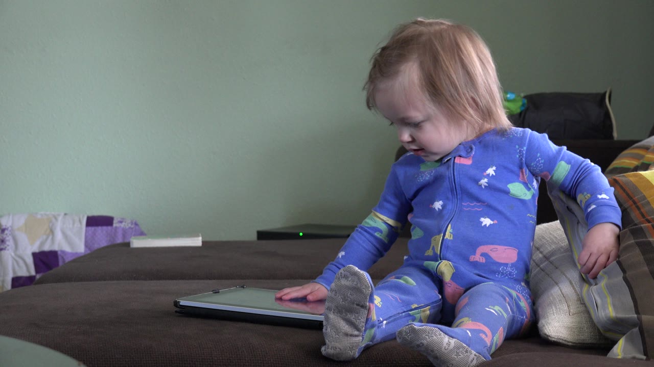 A blonde child in blue pajamas sits on a bed in Westminster, Colorado, looking down while touching a closed gray notebook beside pillows.