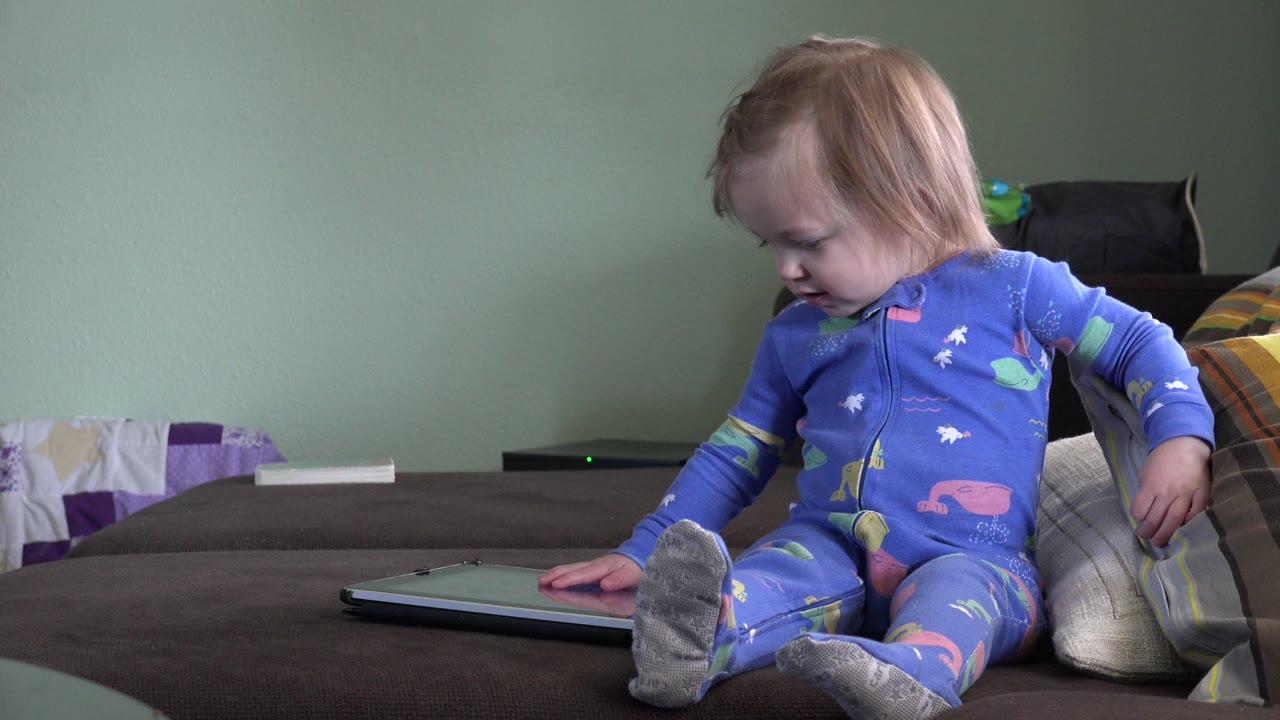 The child remains seated on the bed in Westminster, Colorado, leaning toward the closed gray notebook with pillows behind them.