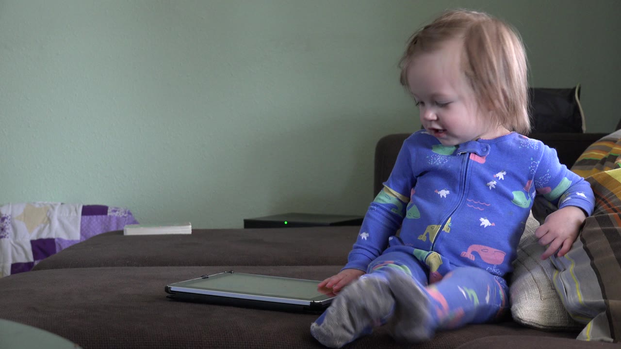 Blonde baby girl in blue pajamas looks down while sitting on a brown sofa near a tablet in a Westminster, Colorado home interior.