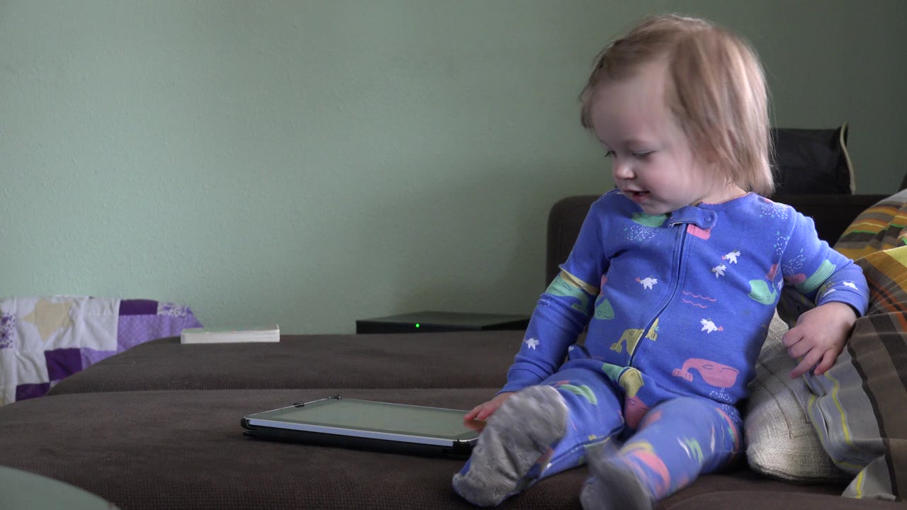 The child remains seated on the sofa in blue pajamas, looking down toward the tablet while pillows and a pale green wall fill the Westminster home background.