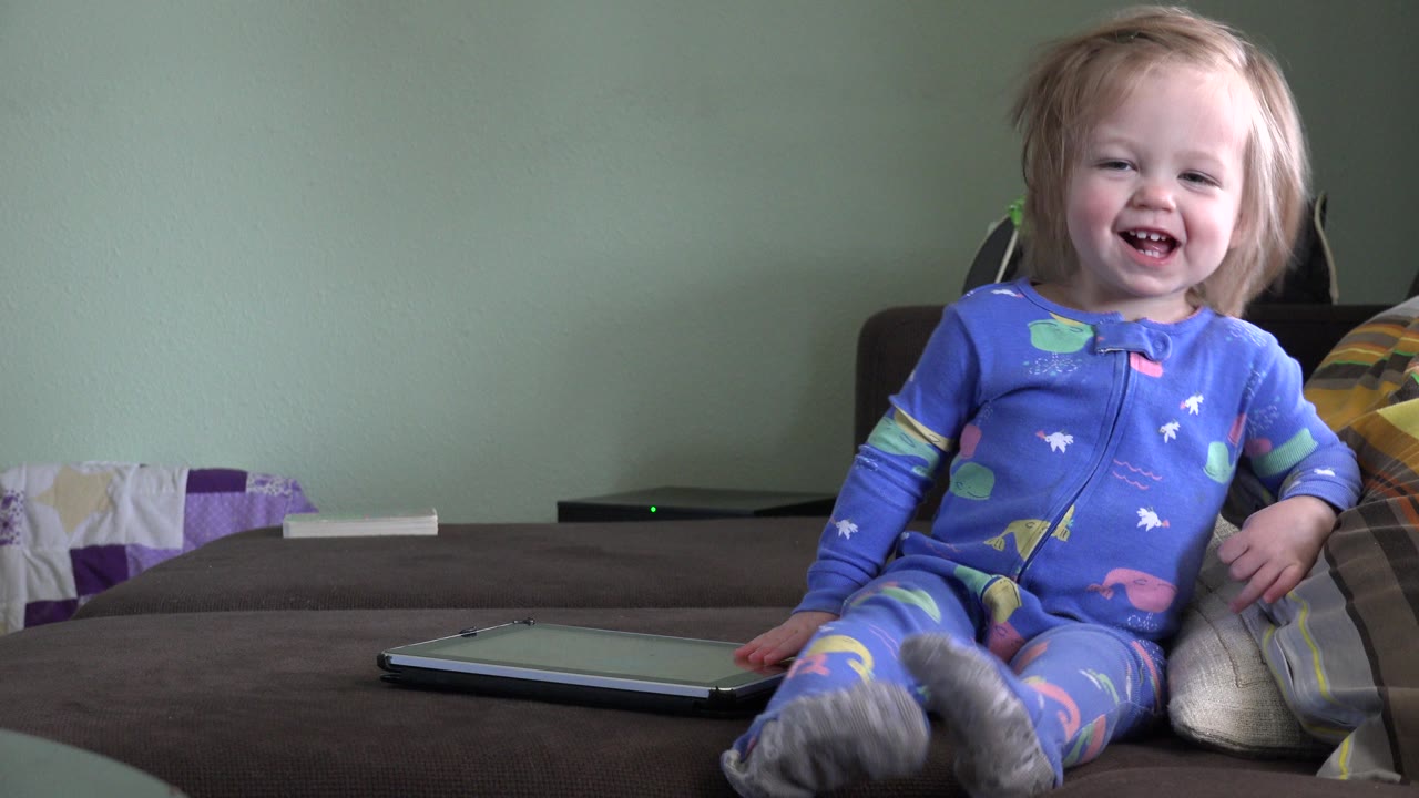 The blonde toddler smiles broadly while sitting on the sofa, with her feet forward, the tablet at lower left, and the Westminster interior wall behind her.