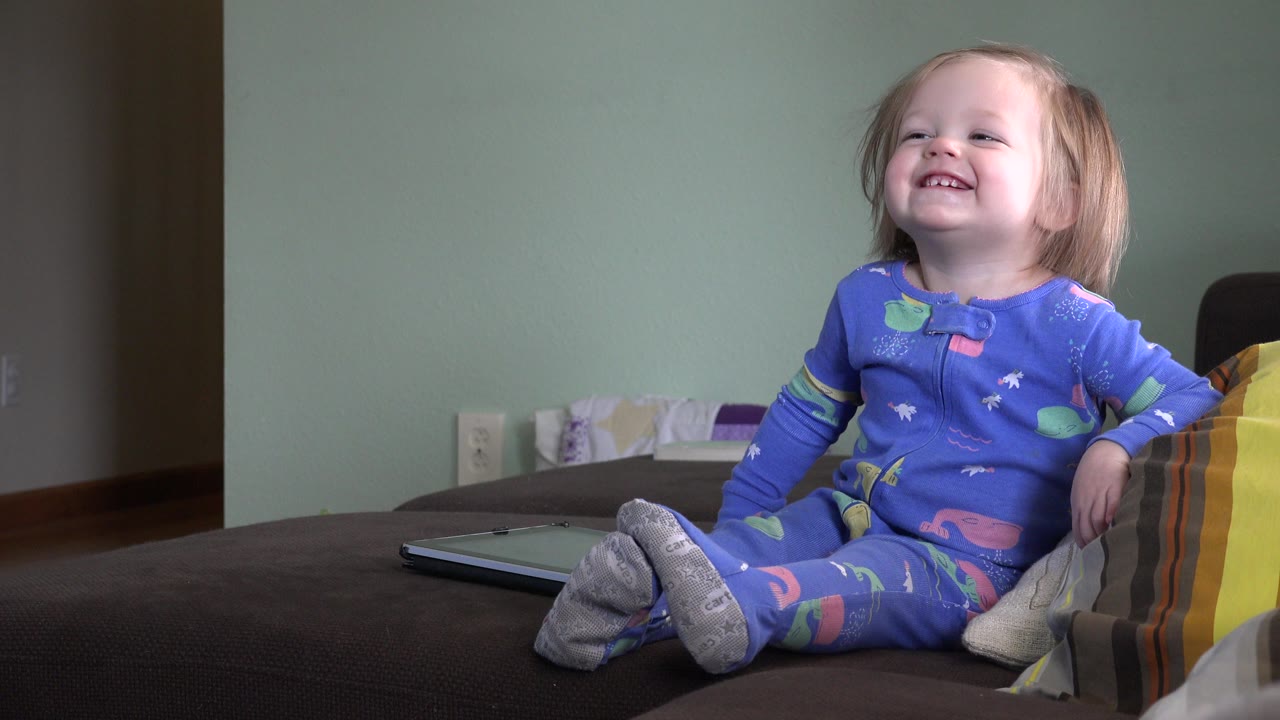 A smiling blonde toddler leans slightly beside striped pillows on a couch inside the Westminster, Colorado home setting.