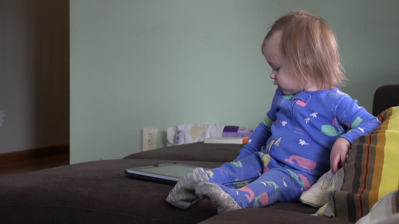 Baby in blue pajamas sitting on a bed and looking down.