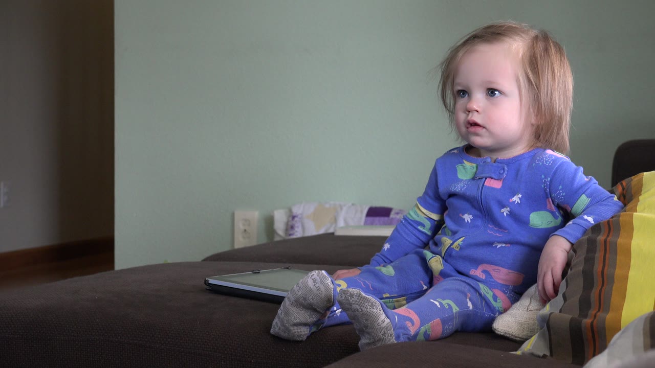 At the start of the Westminster, Colorado clip, a young blonde girl in blue patterned pajamas sits on a dark couch with striped pillows to her right.