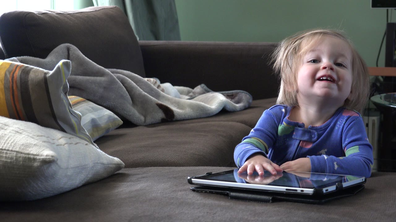 In Westminster, Colorado, a young girl in blue smiles behind a laptop on a low table, with a couch, pillows, and blanket behind her.
