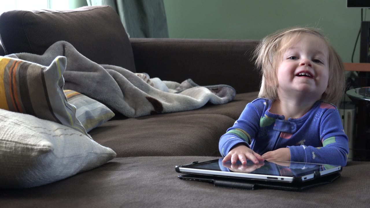 In Westminster, Colorado, the child grins with both hands near the laptop, framed low across the tabletop with household cushions in the background.