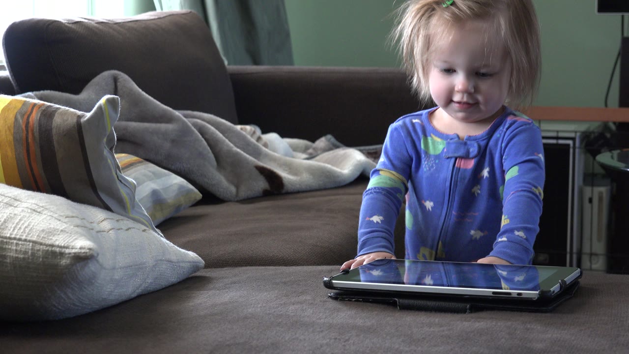 In a Westminster, Colorado living room, a young toddler in blue stands by a couch with both hands near a tablet while pillows and a blanket sit behind her.