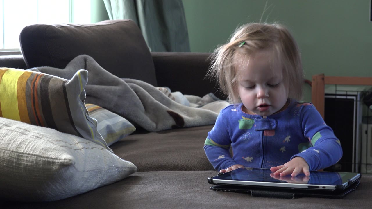 Blonde toddler in blue pajamas touches a tablet on a bed or sofa.