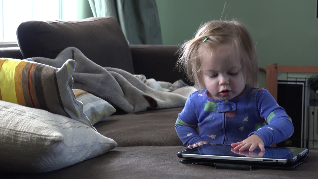 At 0:01 in Westminster, Colorado, the toddler keeps one hand on the tablet while sitting low on the bed or sofa with pillows and blankets behind.
