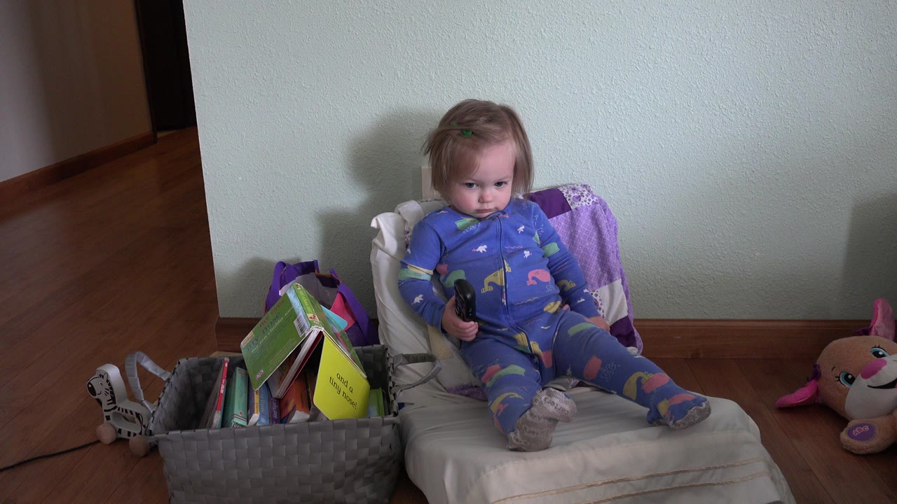Young child sitting on a small cushioned chair beside books and toys.