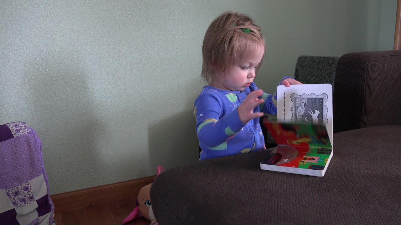 In Westminster, Colorado, a toddler in blue pajamas looks down while opening a colorful board book at a dark table in a home interior.