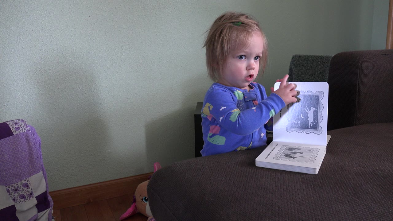 Young child holds an open book behind a brown sofa.