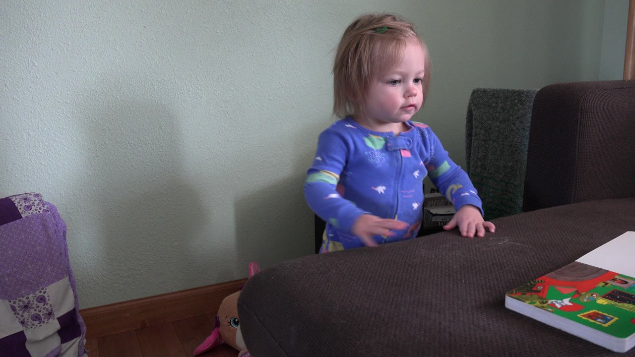 Toddler in blue pajamas stands behind a brown upholstered seat near a book.