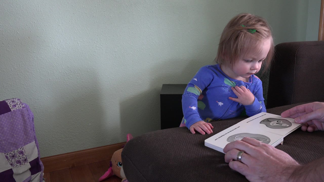 Small child in blue pajamas looking at an open book with adult hands nearby.