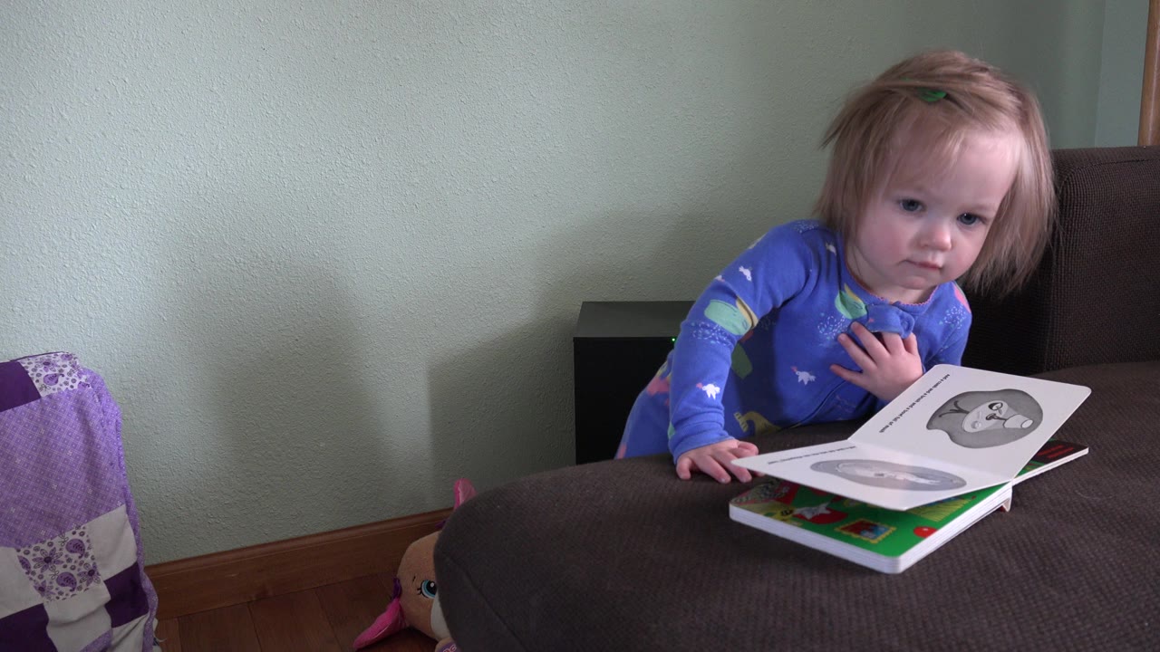 The toddler remains beside the open children's book in the Westminster, Colorado interior, with her hand near her chest and the book spread across the brown cushion.