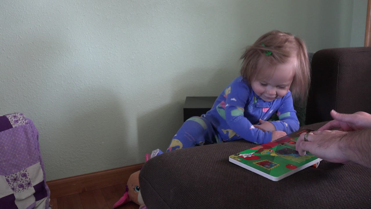 Young girl in blue pajamas looks down at a colorful book beside adult hands.