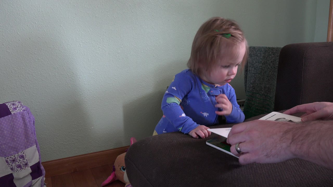 Young child in blue pajamas looking down at a board book beside an adult hand.