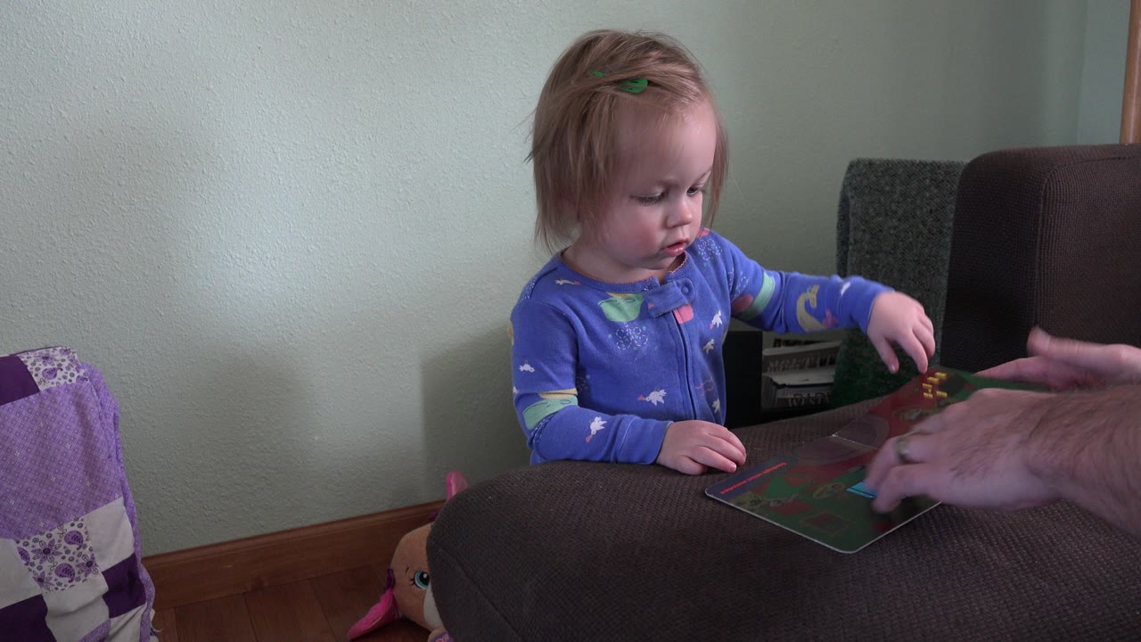 Blond toddler touches an open orange book beside adult hands.