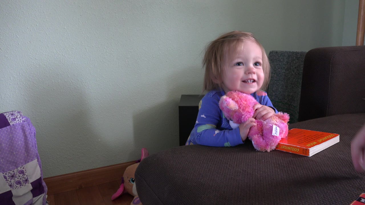 Toddler in blue pajamas holds a pink stuffed animal at a highchair tray.