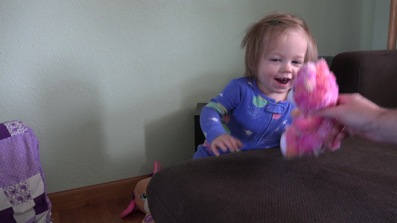 Toddler girl reaching toward a pink plush toy near a brown couch.