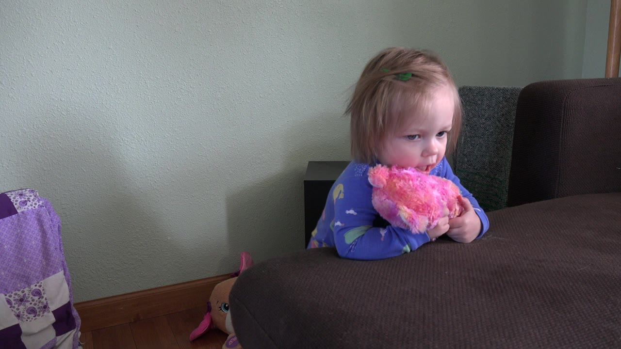 Baby holding a pink plush toy behind a brown sofa.