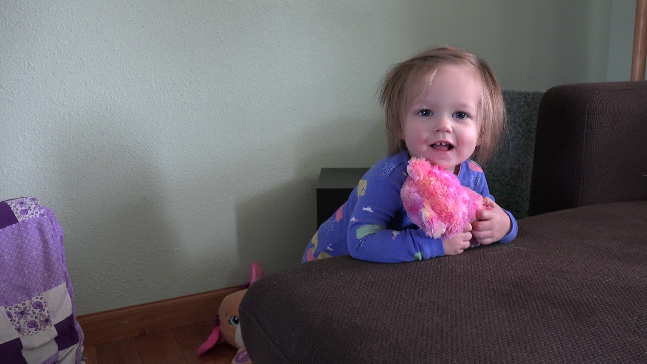 Toddler girl holding a pink plush doll behind a brown bed or sofa.