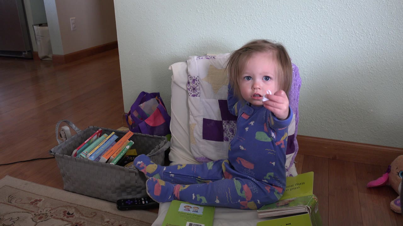 The child remains seated by the book bin in the Westminster living room and extends a hand with a small white object toward the camera.