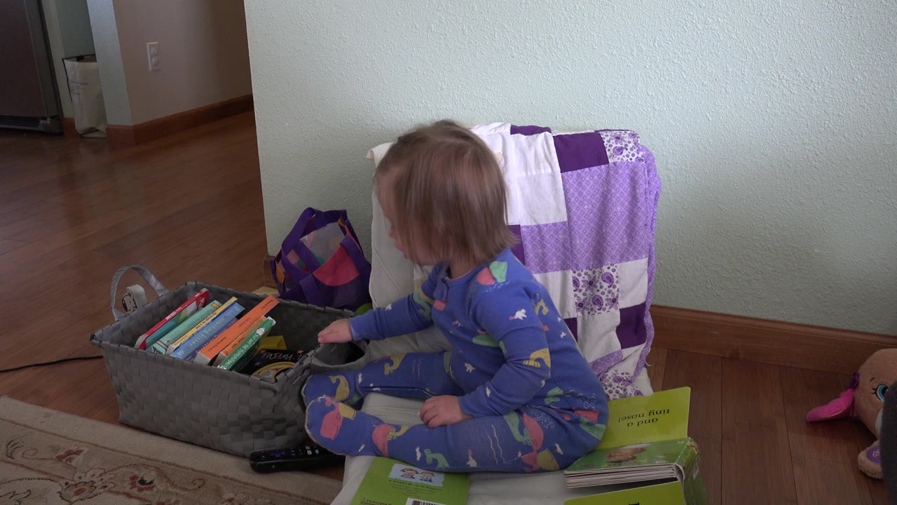 In Westminster, Colorado, a toddler girl in blue pajamas sits sideways on a cushion next to a gray basket filled with children's books and toys.