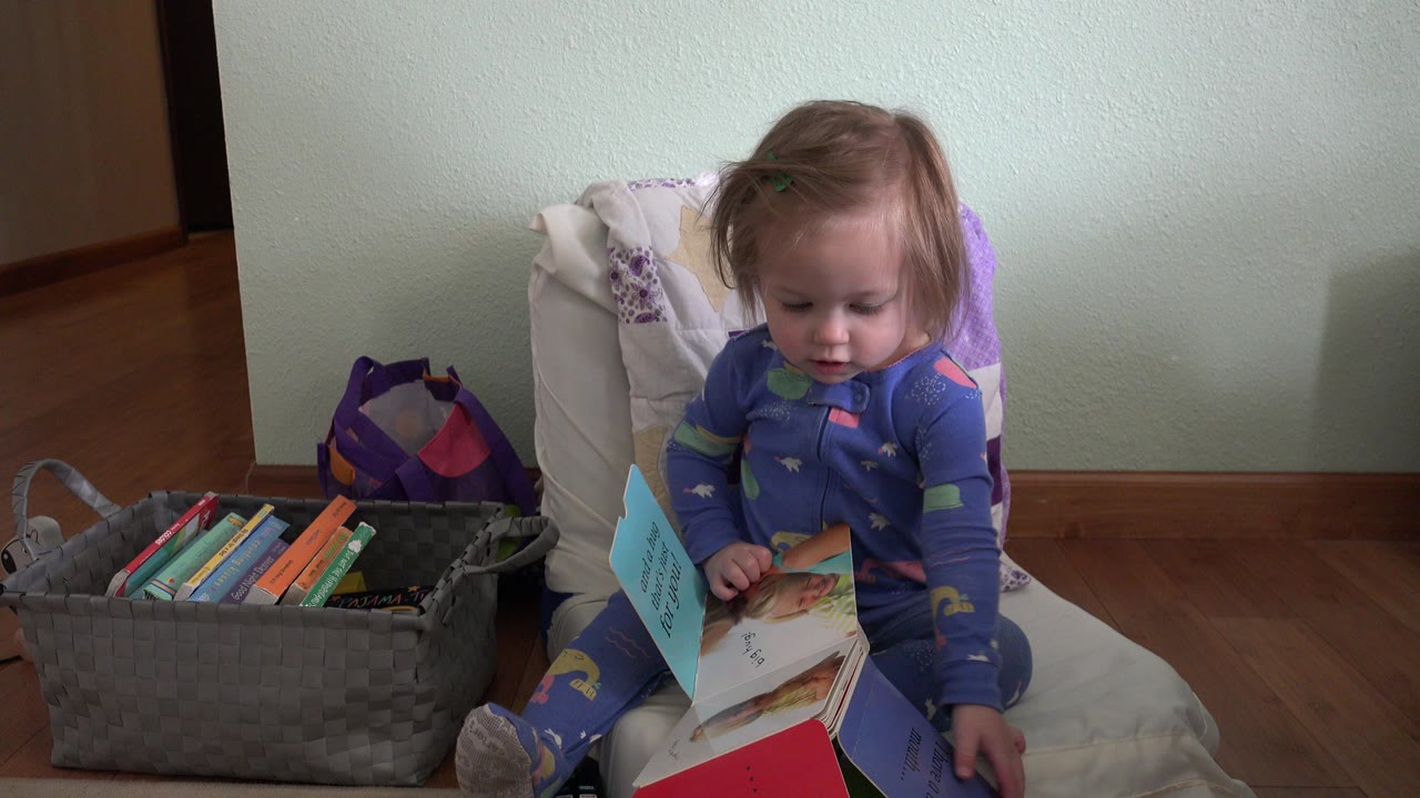 Blonde baby girl sits on a chair with an open children's book.