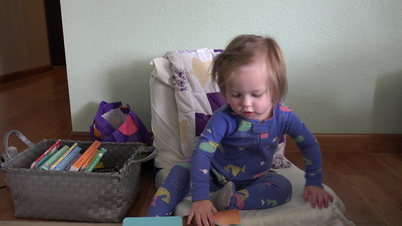 The toddler remains seated in the Westminster home interior, looking down toward books near their lap while the gray book basket stays visible at left.