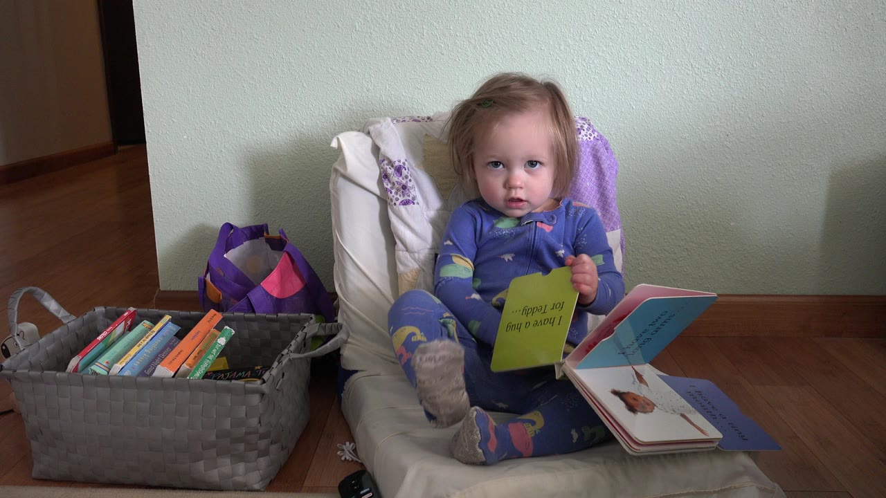 Baby sitting beside a basket of children's books with an open board book in her lap.
