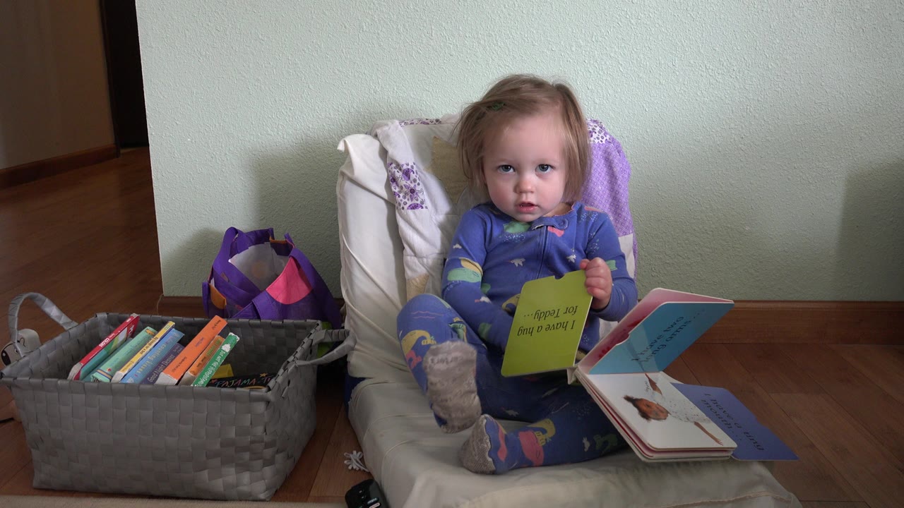 The baby looks forward from the floor chair in Westminster, Colorado, with a green board book in hand, an open picture book on her lap, and a basket of books at her side.