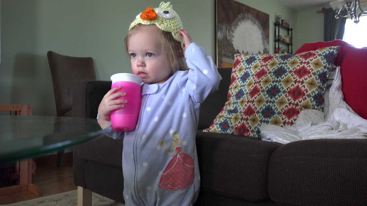 At 0:01, the toddler looks off to the side while keeping one hand on the knitted hat and the other around the pink cup in the living room.