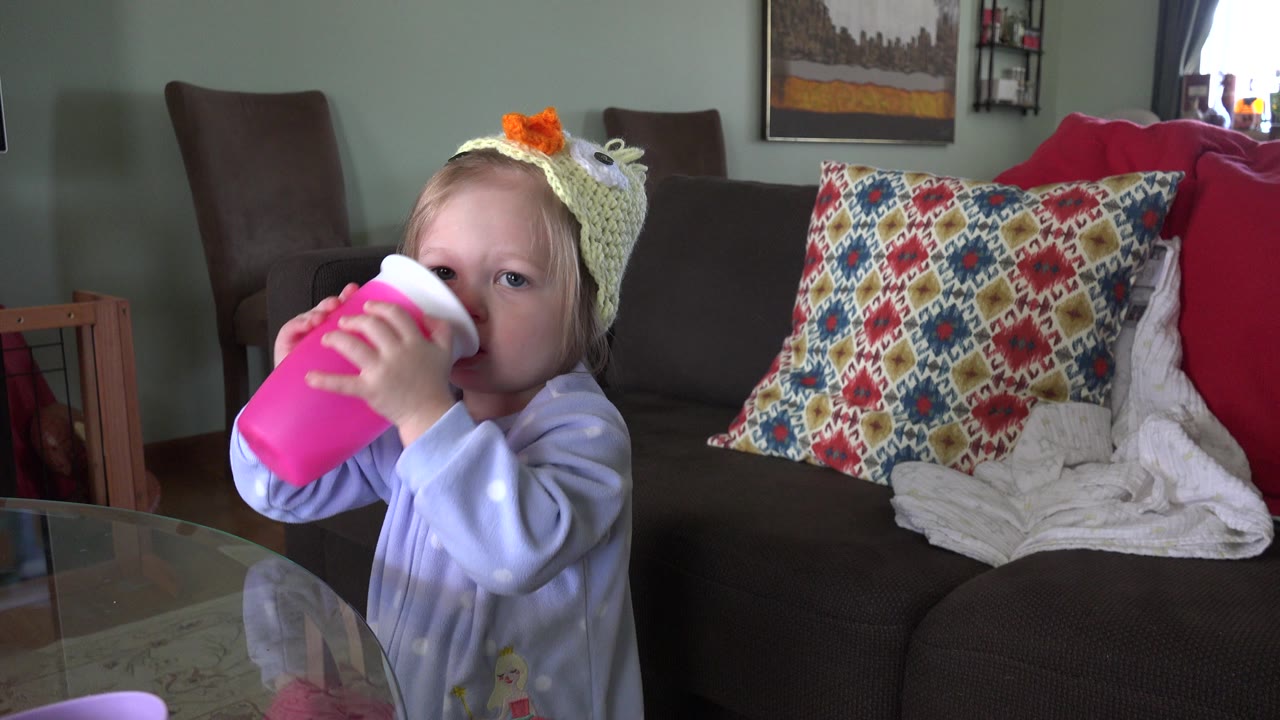 The girl keeps the bright pink cup raised to her mouth in the Westminster, Colorado living room, with her eyes visible above the rim and the couch cushions in the background.