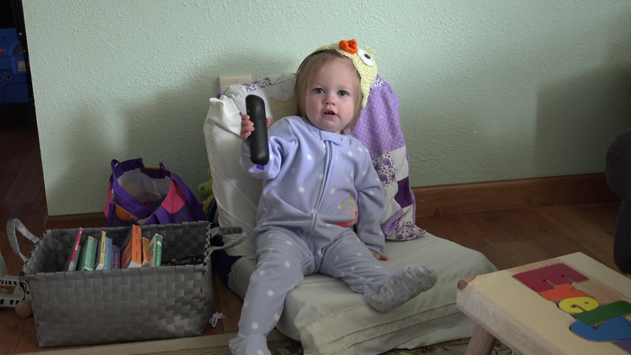 At 0:02 in Westminster, Colorado, the toddler holds the black remote upright toward the camera, with the small table and toy-like colored shapes visible at right.