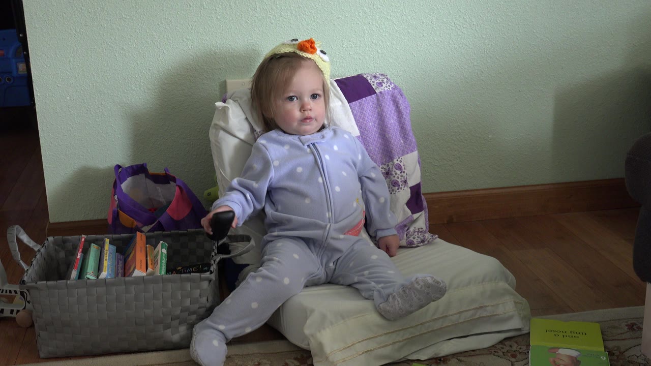 In Westminster, Colorado, a toddler girl sits slightly angled on a cushioned chair beside a woven basket of children's books, with pillows and a green wall behind her.