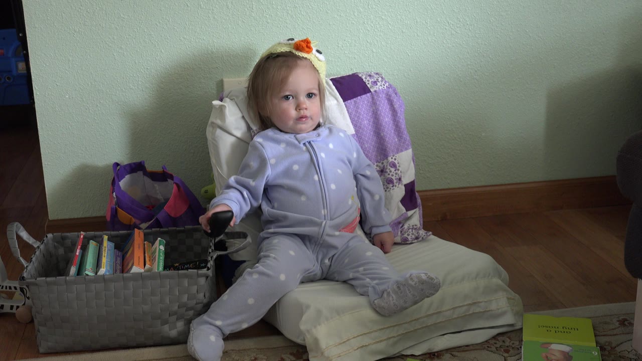 The toddler remains seated in the Westminster home interior, her hand near a dark object at the side of the chair while the children's book basket fills the lower left foreground.