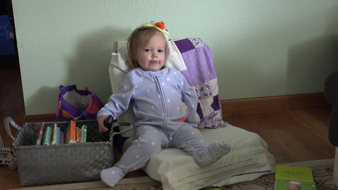 Baby seated on a cushioned chair beside a basket of books and toys.